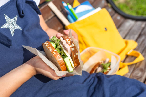 子供の手で昼食。プラスチック容器に入ったサンドイッチ。スナック、学校の朝食、昼食。昼休み。本と学用品の黄色いバックパック - child human hand sandwich lunch box ストックフォトと画像