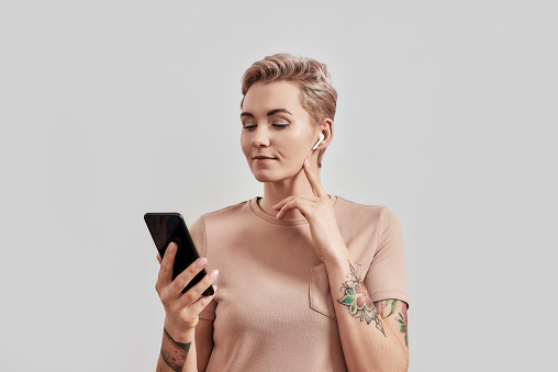 Portrait of tattooed woman with pierced nose and short hair in wireless earbuds or earphones using smartphone isolated over light background. Horizontal shot