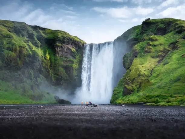 Photo of Skogafoss waterfall, Iceland. Mountain valley and clear sky. Natural landscape in summer season. Icelandic nature. Group of a people near large waterfall. Travel image