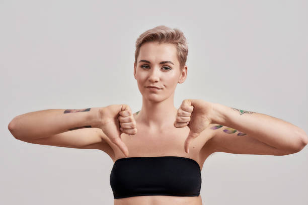 bad. portrait of unhappy half naked tattooed woman with pierced nose and short hair looking at camera, showing thumbs down sign isolated over light background - women tattoo naked sadness imagens e fotografias de stock