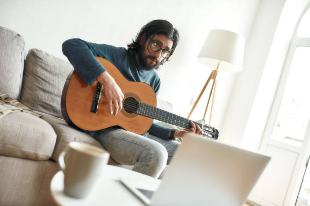 istock Watching video tutorial. Young man sitting on sofa at home and learning guitar online. Sitting on sofa at home and looking at laptop 1257085552