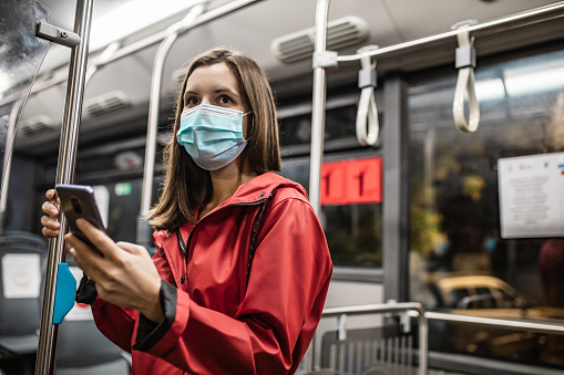 Young woman wearing a protective mask while sitting in a bus and using a mobile phone