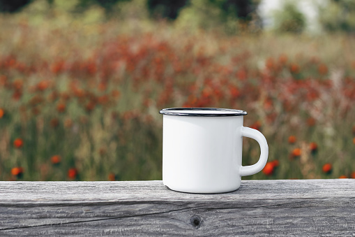 Close up of metal mug on old wooden table, board with defocused blooming mountain meadow. Outdoor tea, coffee time. Mockup of white enamel cup, lifestyle relax, trekking and camping concept.