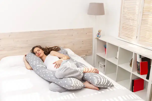 Photo of Beautiful sleeping smiling pregnant woman with a big tummy abdomen with long curly hair and closed eyes, hugging a long body pillow with her arm and legs and lying on the bed indoor.