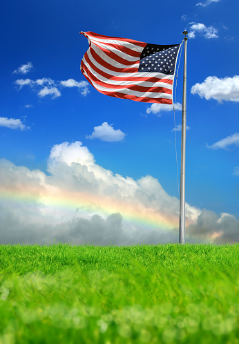 Conceptual image of waving American flag at flagpole on green landscape over sunny sky with rainbow