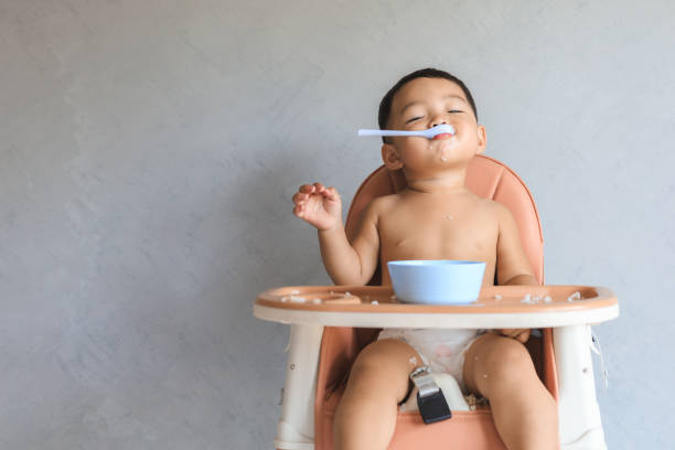 Asian baby boy eating food by himself Happy infant Asian baby boy eating food by himself on baby high chair and making mess with copy space. infant feeding stock pictures, royalty-free photos & images