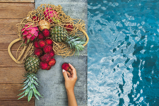 Woman hand hold rambutan from heap fresh tropical fruits pineapple, rambutan, dragon fruit in reusable textile bag on pool edge. Raw healthy food and vegetarian diet. Zero waste.