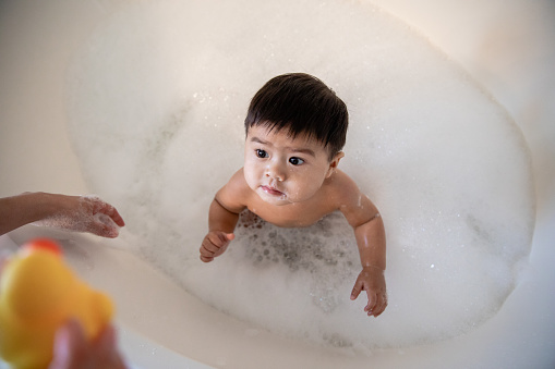 Happy boy splashes in bubble bath while mother holds rubber duck