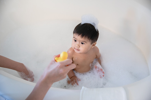 Mother hands rubber duck to adorable baby boy in bubble bath
