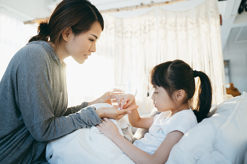 Young Asian mother holding a glass of water and giving medicine to sick little daughter lying in bed