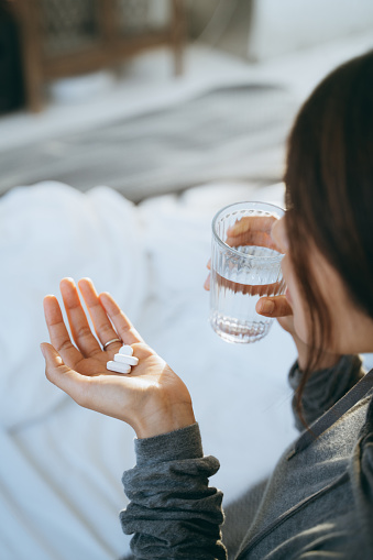 Cropped shot of young Asian woman lying in bed and feeling sick, taking medicines in hand with a glass of water