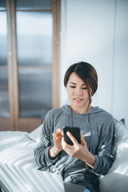 istock Young Asian woman lying in bed using smartphone and working on laptop on her lap, working from home in the morning 1257145570