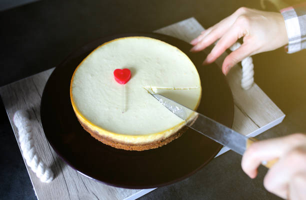 Girl is cutting with a knife classical vanilla New York cheesecake on dark-violet plate on wooden tray with rope handles. Small red heart is lying on dessert. Perfect hygge Valentine's day present.
