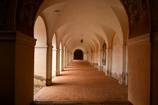 View of an interior of an old monastery with arches along the walls, old-fashioned religious decorations, brick-based floor and some sources of light hanging from the ceiling seen in Poland in summer