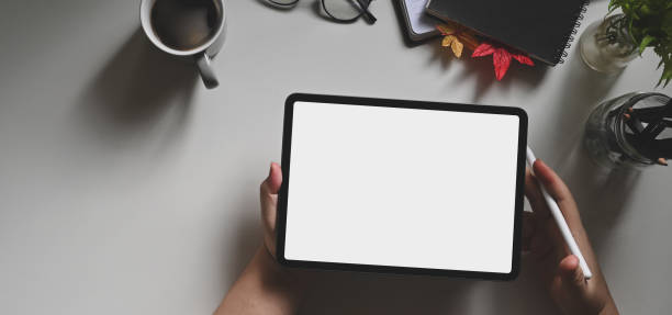 Workspace hands are holding a white blank screen tablet with a stylus pen at the white working desk.