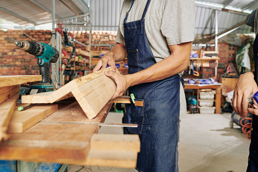 Cropped image of carpenter in denim apron making wooden furniture in workshop