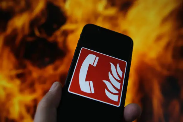 Photo of View on hand holding mobile phone with international fire emergency telephone symbol. Blurred fire flames background.