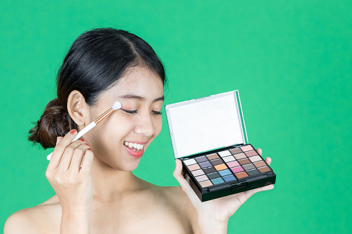 Healthy and cosmetics concept. Beauty face of young Asian woman applying make up with brush over green isolated background.