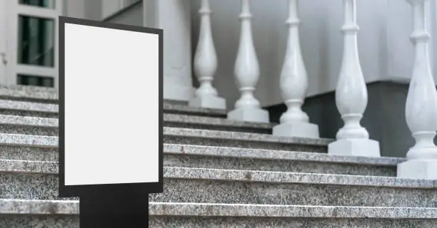 empty screen in black metal frame with white place for mock-up stands on steps of stone staircase near entrance to house or hotel outside