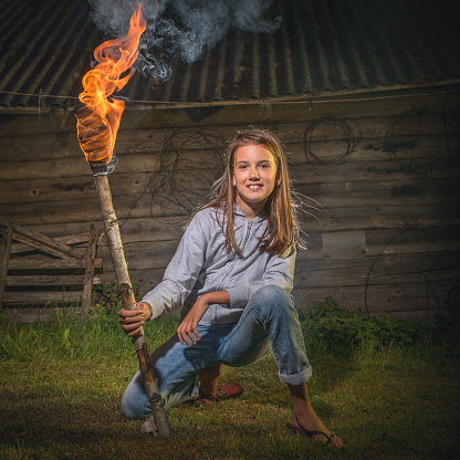 A girl with a burning torch sits near the barn at night.