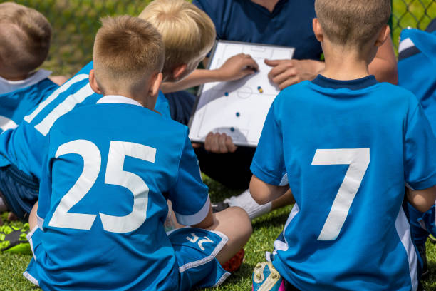 grupa młodych chłopców w niebieskich koszulkach siedząc na sports grass field witch school coach. dzieci słuchanie trener's tactic talk. młody trener wyjaśnia taktykę piłki nożnej. coaching młodych zawodników w sporcie - sports uniform blue team event sports activity zdjęcia i obrazy z banku zdjęć