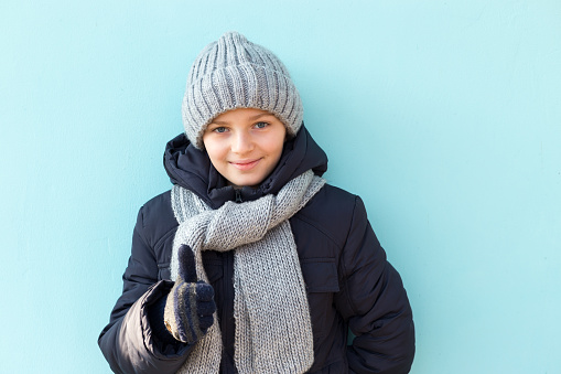 Funny smiling child showing thumbs up, ready for winter vacation. Fashionable boy in winter gray cap and scarf standing against blue wall.