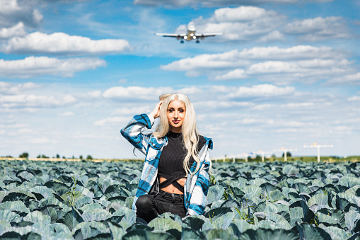 Fashionable dyed blonde transgender man with feminine styling, make-up and outfit  standing outdoors in agriculture cabbage field close to the airport. Passenger Airplane landing in the background. (No edit - real shot). Colorful vibrant Surreal Transgender Female Fashion Lifestyle Portrait