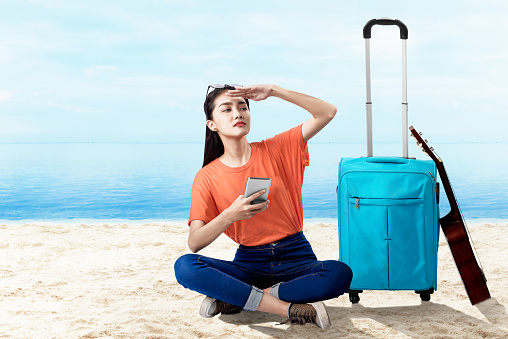 Asian woman sitting with a suitcase holding mobile phone looking at scenery view on the beach with a blue sky background