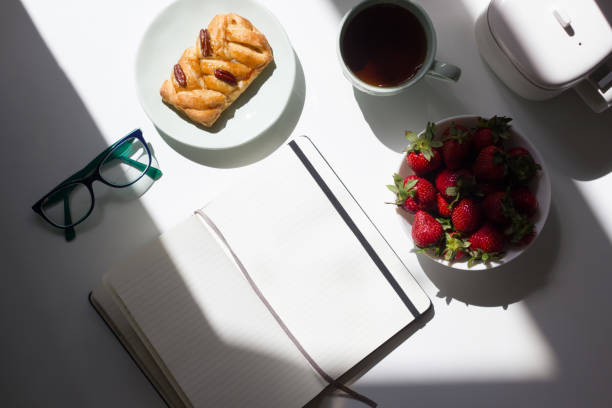 Blank open notepad with glasses, cup of tea, tea pot and strawberries in home office on white background