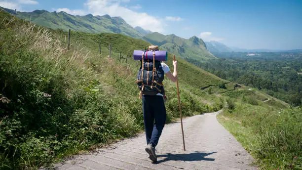 Young Caucasian man with straw hat, stick and backpack, on a path between mountains and a village on the horizon