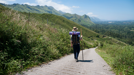 Young Caucasian man with straw hat, stick and backpack, on a path between mountains and a village on the horizon