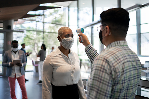 Receptionist measuring temperature of female employee at office's entrance - with face mask