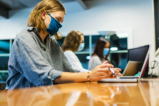 Three female college students; One in foreground in focus two female and one male blurred in background wearing face masks using technology working on laptops part of a series (Shot with Canon 5DS 50.6mp photos professionally retouched - Lightroom / Photoshop - original size 5792 x 8688 downsampled as needed for clarity and select focus used for dramatic effect)