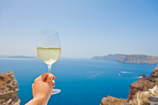 Female hand holding a glass of cool white wine on a hot summer day with a view of the aegean sea.