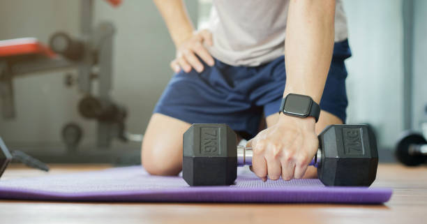 close up young man with smart watch holding dumbbell and sitting over exercise mat on the floor while resting after workout at home for new normal concept close up young man with smart watch holding dumbbell and sitting over exercise mat on the floor while resting after workout at home for new normal concept fitness tracker stock pictures, royalty-free photos & images