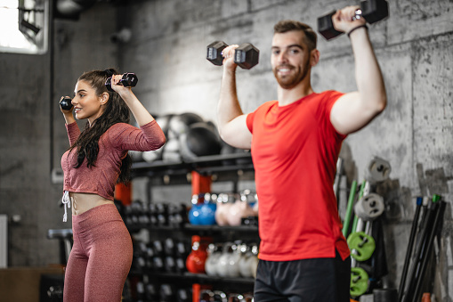 Young sporty man is having weight training with dumbbells at the gym with the assistance of young female fitness instructor. Focus on woman
