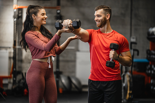 Young sporty man is having weight training with dumbbells at the gym with the assistance of young female fitness instructor.