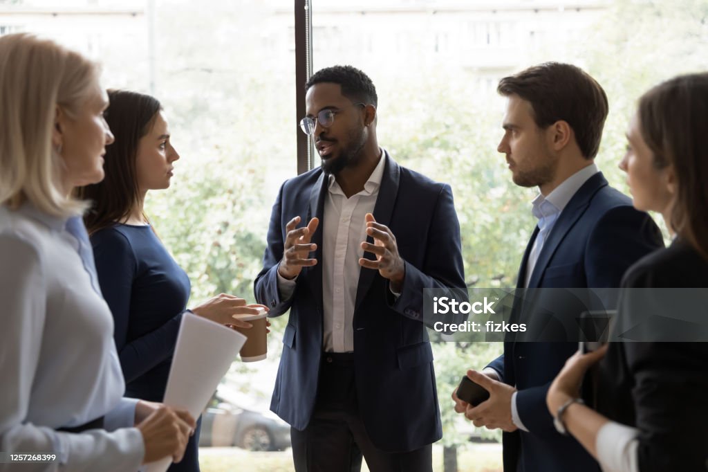 Confident African male leader telling diverse colleagues about new project Confident African male leader standing telling diverse colleagues about new project, boss lead briefing for different age workgroup team employees in office. Sharing information and leadership concept Business Stock Photo