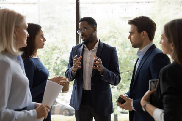 Confident African male leader telling diverse colleagues about new project Confident African male leader standing telling diverse colleagues about new project, boss lead briefing for different age workgroup team employees in office. Sharing information and leadership concept persuasion stock pictures, royalty-free photos & images