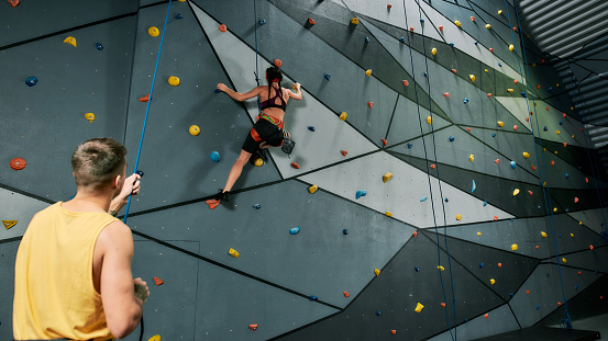 Male instructor holding rope, teaching, looking at woman in safety equipment and harness while she is training on the artificial climbing wall. Bouldering training concept. Web Banner