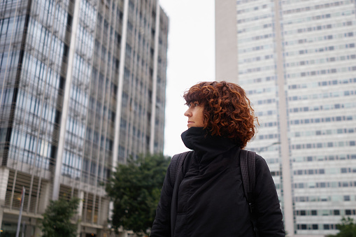 Young attractive woman in black casual wear and curly hair standing with skyscrapers and big city background looking to the left side. Big city and traveling concept.