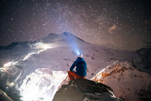 He looks up to a starry sky, shining his headlamp toward snowy mountain range