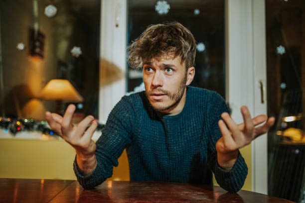 Confused man Portrait of young attractive confused man explaining and gesturing while sitting at the table in night in Christmas decorating room confusion raised eyebrows human face men stock pictures, royalty-free photos & images