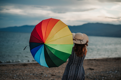 Beautiful young woman with rainbow umbrella on windy summer day by the sea