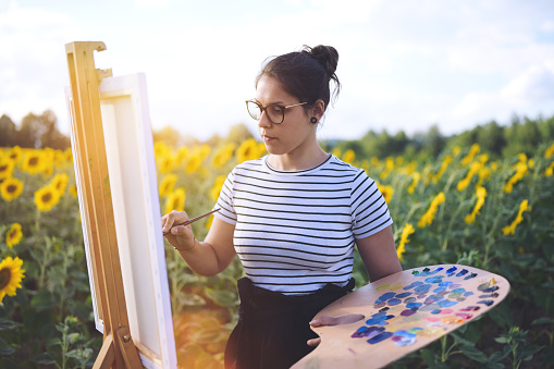 young female artist painting outdoors in sunflower fields holding a big artistic pallet and mixing colors