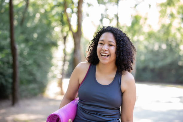 женщина улыбается, держа коврик йоги и ходить на открытом воздухе для физичес�ких упражнений - stretching yoga exercise mat women стоковые фото и изображения