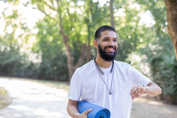 istock Man checks fitness tracker while holding exercise mat and walking in park for exercise 1257301478