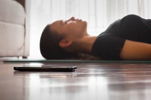 A woman lying on a yoga mat at home with her eyes closed listening to a yoga guide app and music.