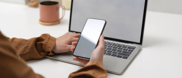 istock Female hands holding mock up smartphone while sitting at worktable with mock up laptop in home office 1257319195