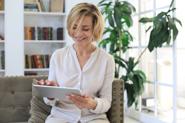 istock Cheerful middle aged woman sitting on sofa, using computer tablet apps, looking at screen,reading good news in social network, shopping or chatting online. 1257337360